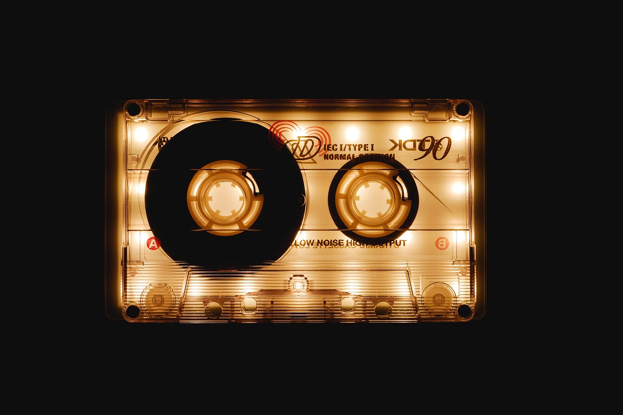Une cassette audio transparente posee sur fond noir et eclairee par-derriere, la lumiere doree traversant le boitier, les mentions IEC I/TYPE I NORMAL POSITION et LOW NOISE HIGH OUTPUT lisibles, les reperes A et B en rouge