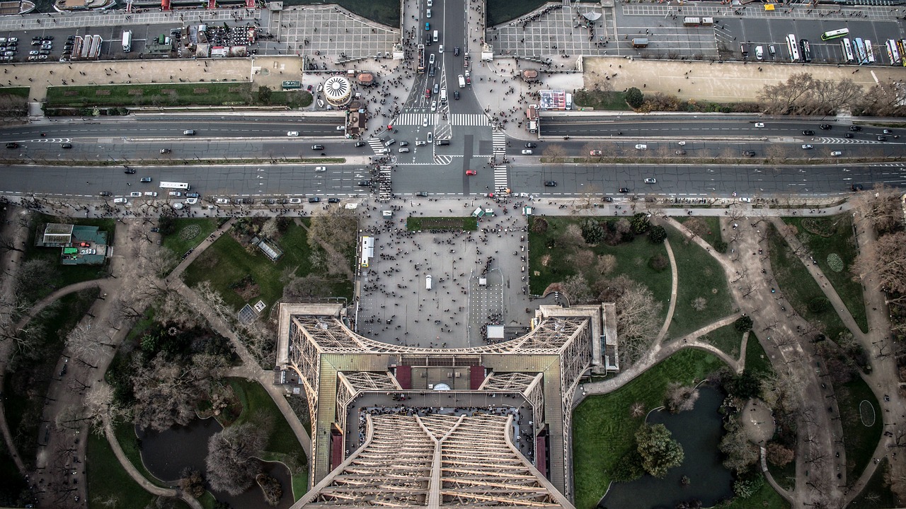 Vue aerienne prise a la verticale depuis le sommet de la tour Eiffel: les quatre piliers et le treillis metallique occupent le bas du cadre, au-dessus les jardins, les allees, un large carrefour et des passages pietons ou l'on distingue la foule
