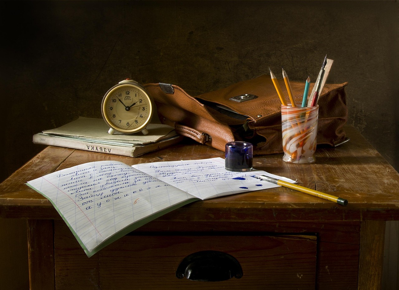 Nature morte sur un pupitre de bois: un cahier ouvert couvert d'une ecriture manuscrite EN CYRILLIQUE avec deux taches d'encre bleue, un crayon jaune, un encrier de verre, un pot de crayons, une sacoche de cuir brun et un reveil creme pose sur des livres