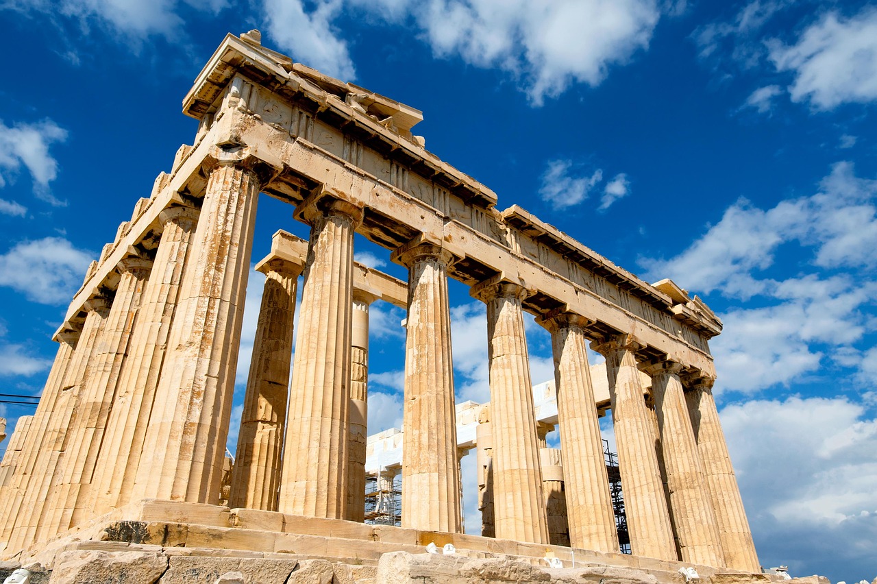 Le Parthenon photographie d'en bas et de biais, colonnes doriques et entablement couleur miel se detachant sur un ciel bleu franc parseme de nuages blancs, avec un echafaudage metallique visible au pied de l'edifice a droite